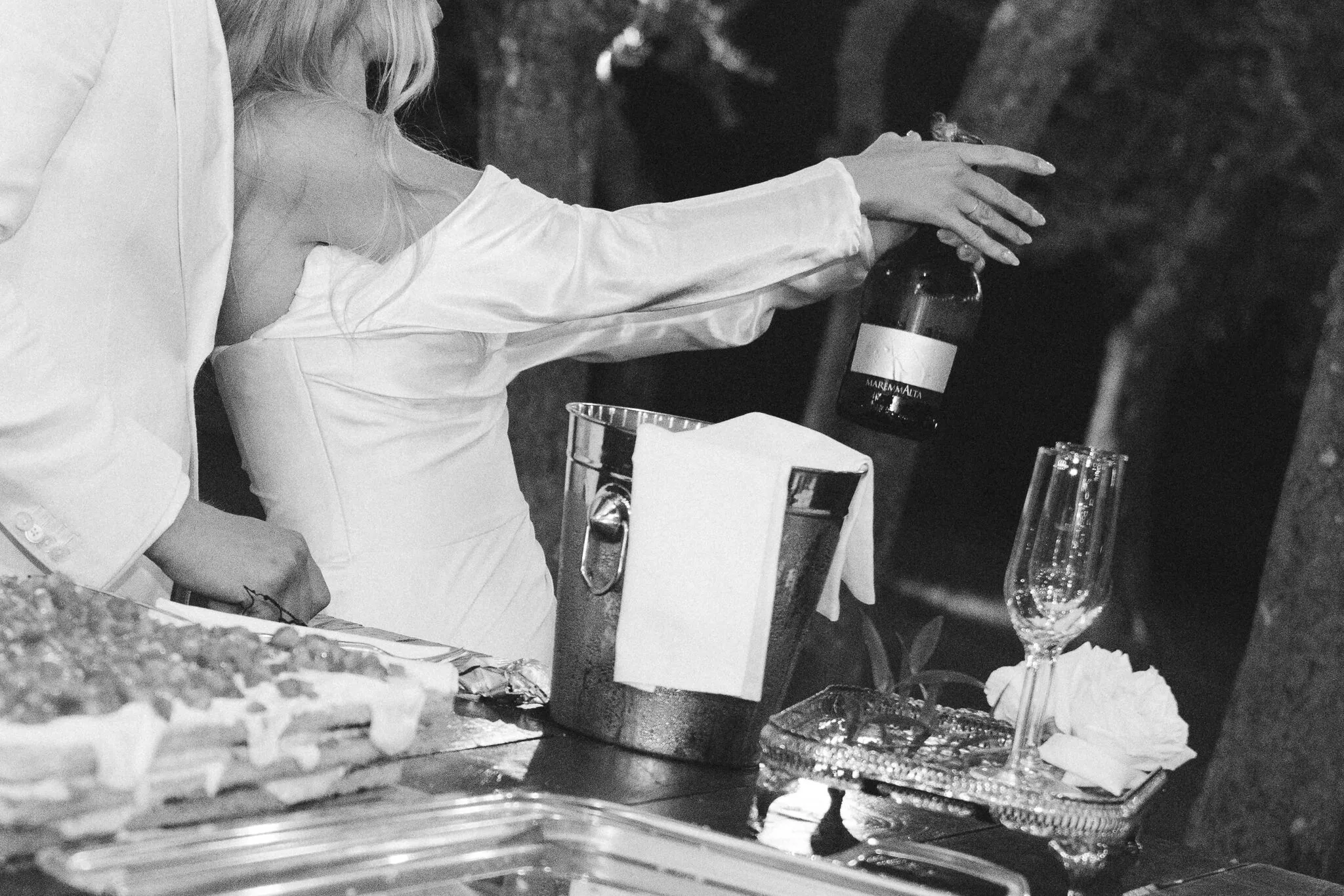 Black and white photo of a bride reaching for a champagne bottle in an ice bucket at an evening celebration