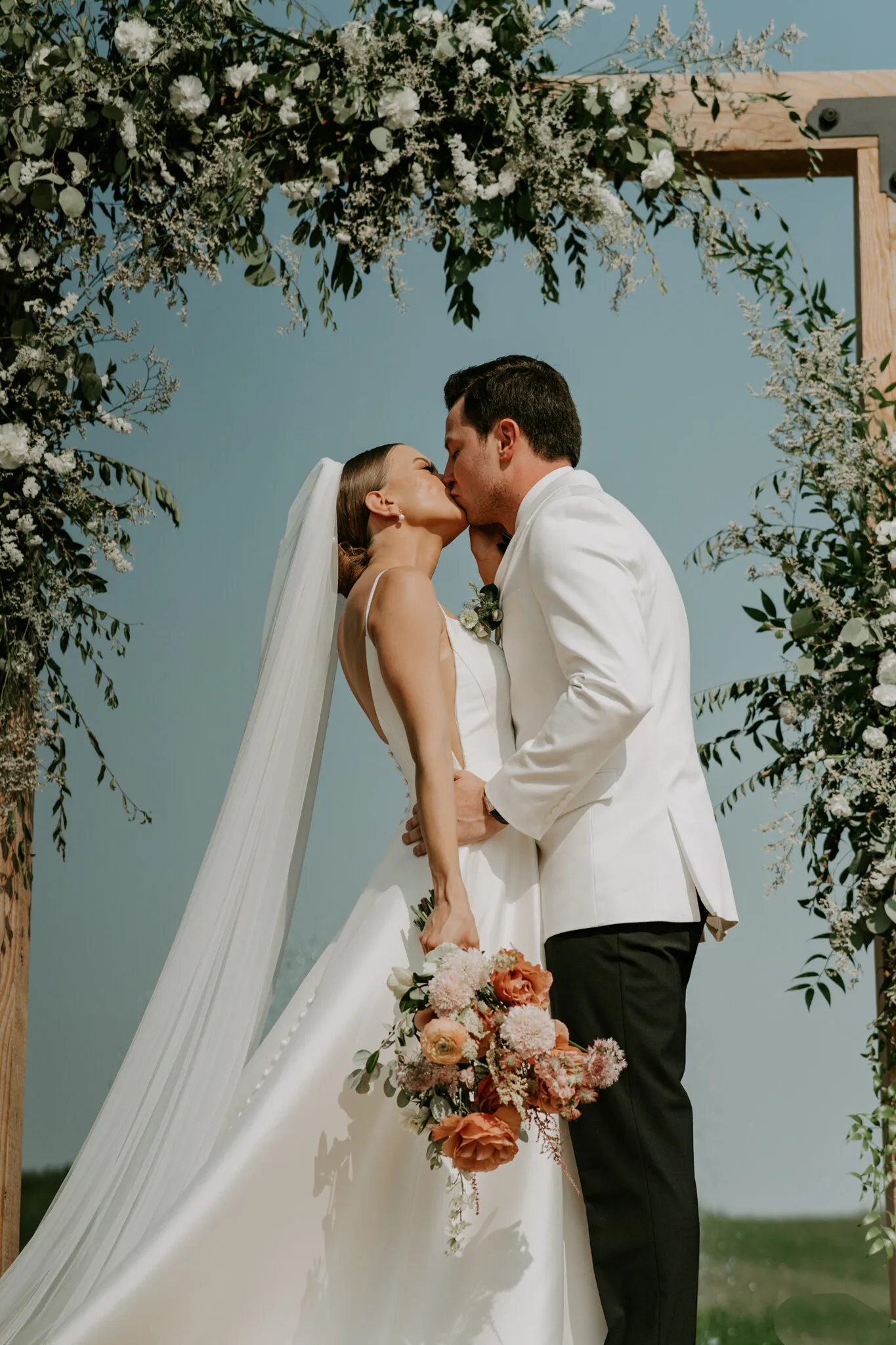 Bride and groom kissing under a wooden ceremony arch covered in white flowers and greenery
